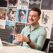 A man smiling and gesturing with his hand in front of a wall decorated with colourful music and album covers, seated at a white table with a laptop.