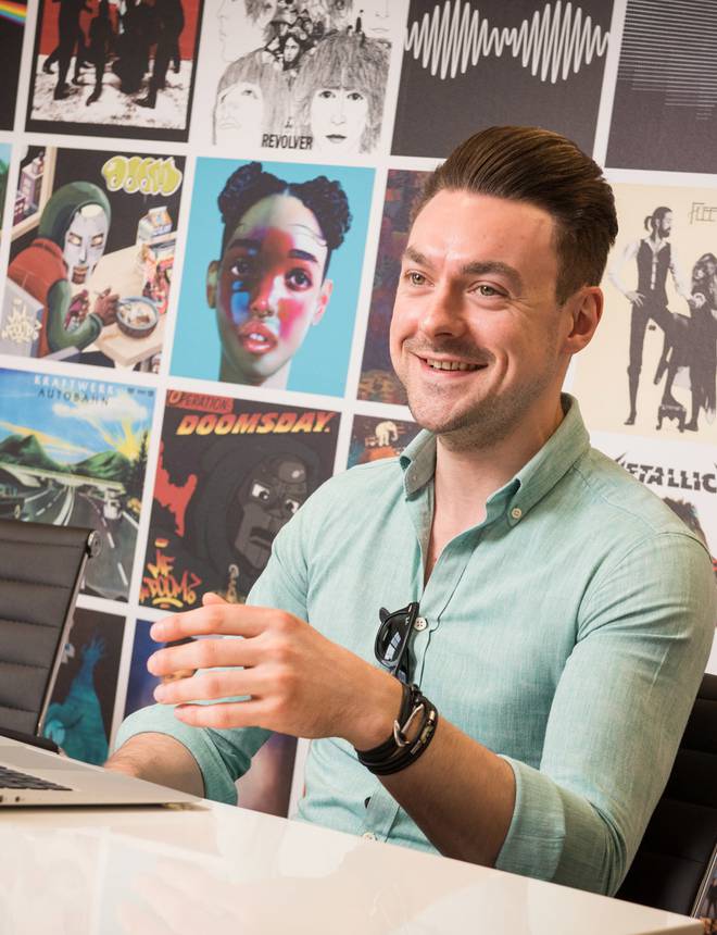 A man smiling and gesturing with his hand in front of a wall decorated with colourful music and album covers, seated at a white table with a laptop.