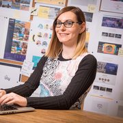A woman with shoulder-length blonde hair, glasses, and a patterned top, sitting at a wooden desk with a wall covered in posters and notes behind her.