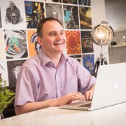A man in a pink shirt smiling while working on a white laptop at a desk, with a collage of colourful posters on the wall behind him.