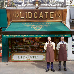 Two smiling butlers stand outside a butcher shop under a green awning, with a wooden sign reading "Lidgate" and a glass display filled with meat products.