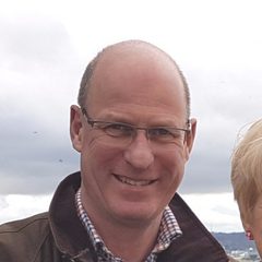A smiling man with glasses, wearing a checkered shirt and dark jacket, outdoors against a grey cloudy sky, with part of a woman's head visible on the right.