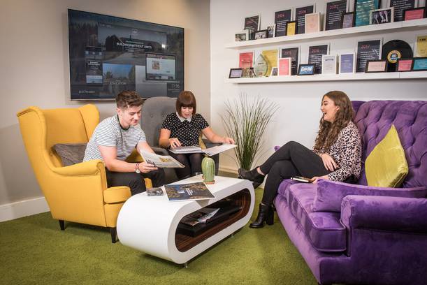Three young women seated on colourful sofas in a conversation, with magazines on the coffee table, in a modern lounge with framed awards on the wall.